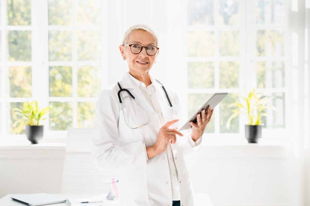 Doctor using digital tablet at hospital, looking at camera in white coat with stethoscope