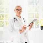Doctor using digital tablet at hospital, looking at camera in white coat with stethoscope